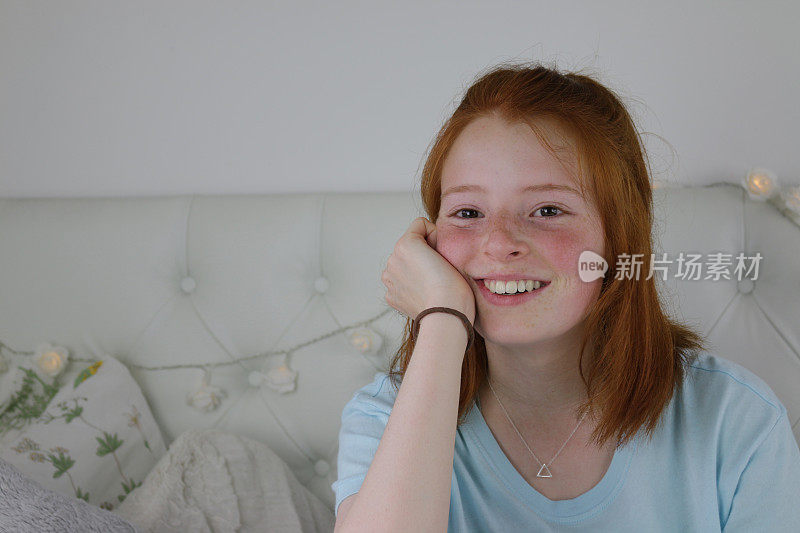 这是一个14 / 15岁的红发少女，皮肤苍白，脸上有雀斑，脸颊通红，坐在卧室里，看起来很开心，手托着下巴微笑着